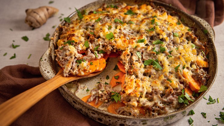 Auflaufform mit überbackener Hackfleisch-Karotten-Schicht, belegt mit Petersilie, daneben ein Holzlöffel und brauner Stoff.