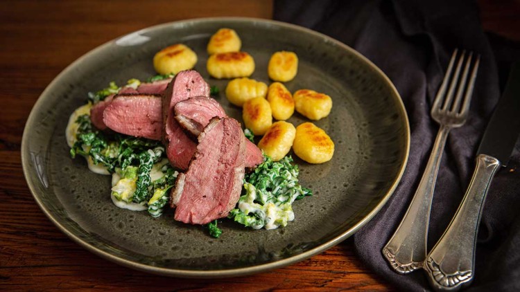 Grauer Teller mit rosa gebratenem Fleisch, cremigem Rahmwirsing, goldbraunen Gnocchi, auf Holztisch mit Besteck rechts.