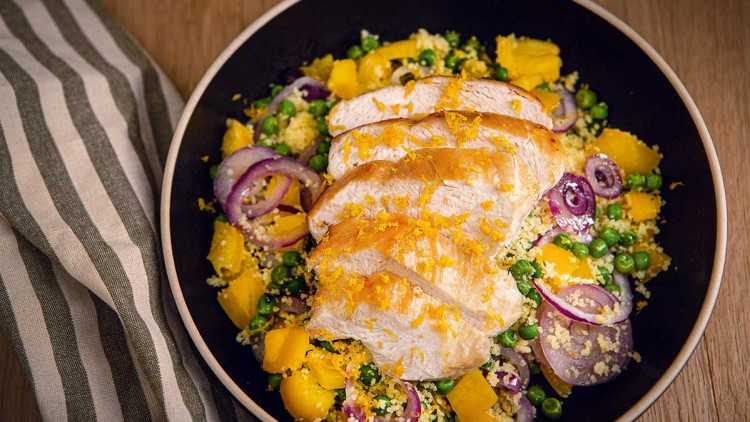 Gebratene Hähnchenbrust auf Couscous mit gelbem Gemüse, Erbsen und roten Zwiebeln in einer Schüssel, daneben ein gestreiftes Tuch.