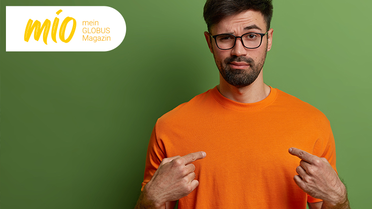 Person in orangefarbenem T-Shirt vor grünem Hintergrund, zeigt mit beiden Händen auf das T-Shirt, Text: mio mein GLOBUS Magazin.