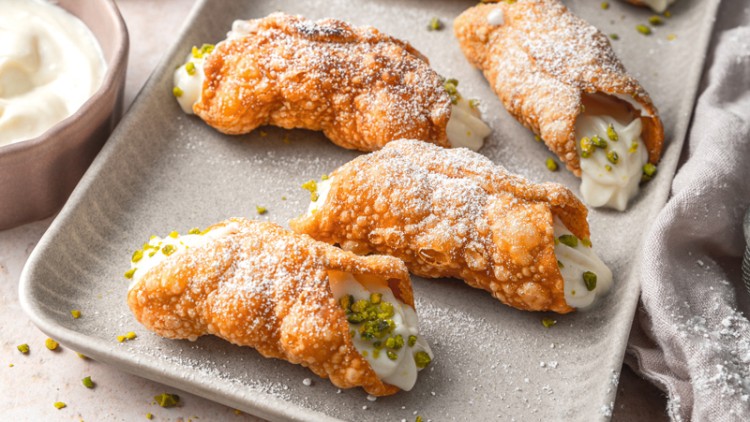 Gefüllte Cannoli mit Puderzucker und gehackten Pistazien auf einem rechteckigen Teller, daneben eine Schale mit weißer Creme.