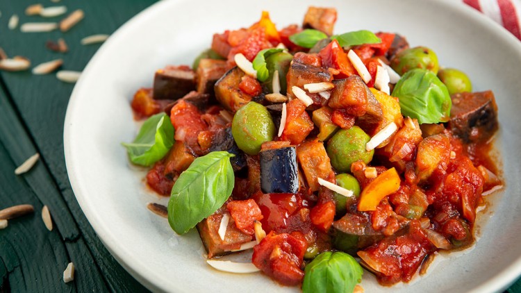 Teller mit Ratatouille aus Auberginen, Tomaten, Paprika und Oliven, garniert mit Basilikumblättern und Mandelsplittern.