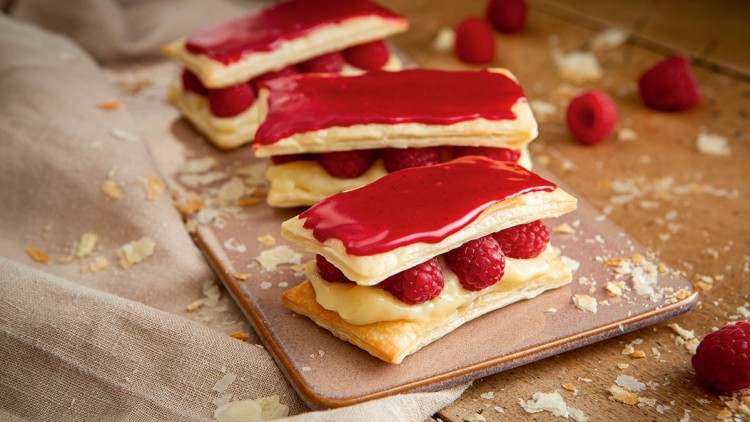 Blätterteig mit gelber Creme, frischen Himbeeren und rotem Überzug auf braunem Teller, umgeben von Krümeln und Stoff.