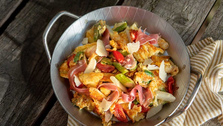 Metallschale mit gebratenem Gemüse, Tomaten, grünem Spargel, Schinkenstreifen und Parmesan auf einem Holztisch mit Stoffserviette.