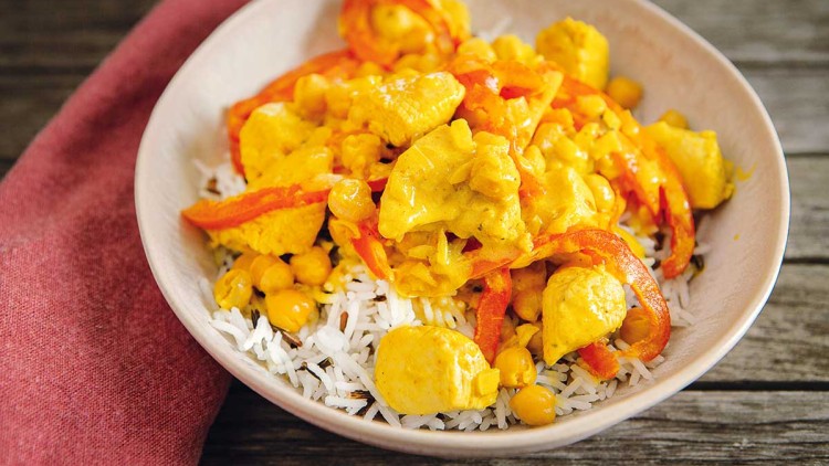 Eine Schale mit Reis, darauf gelbes Curry mit Hähnchenstücken, Kichererbsen und roten Paprikastreifen, daneben ein rotes Tuch.