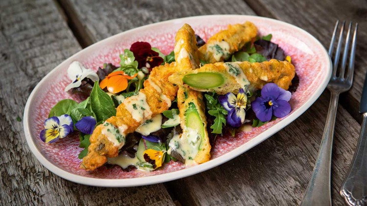 Knusprig frittierter grüner Spargel auf gemischtem Salat mit essbaren Blüten und heller Soße, serviert auf rosafarbenem Teller.