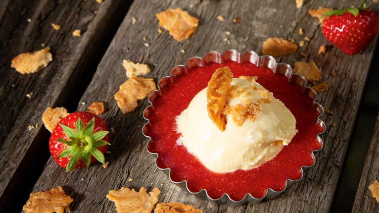 Zwei kleine Metallförmchen mit roter Erdbeersoße, Vanilleeiskugel und knusprigen Stückchen, daneben frische Erdbeeren.