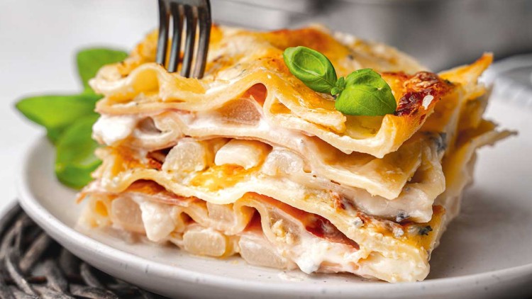 Ein Stück Lasagne mit goldbrauner Kruste und Basilikum auf weißem Teller, eine Gabel wird in die Lasagne gesteckt.