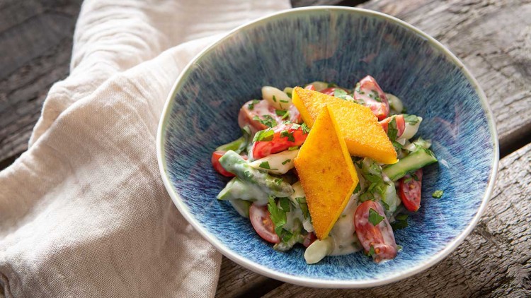 Eine blaue Schale mit Salat aus Tomaten, Gurken, Kräutern und Joghurtdressing, dekoriert mit goldgelben Polentastücken.