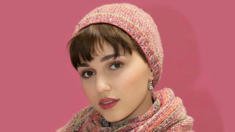 Person trägt ein gestricktes rosafarbenes Set aus Mütze und Schal vor einem einfarbig rosa Hintergrund.