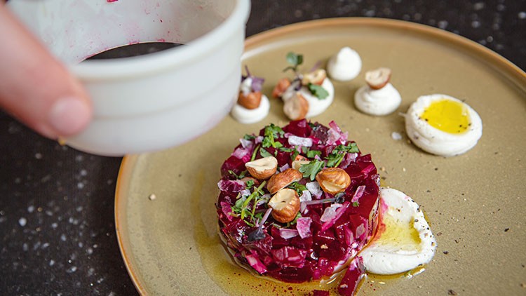 Ein Teller mit Rote-Bete-Tatar, Haselnüssen, Cremehäufchen und Kräutern, daneben wird dunkle Sauce aus einem Gefäß gegossen.