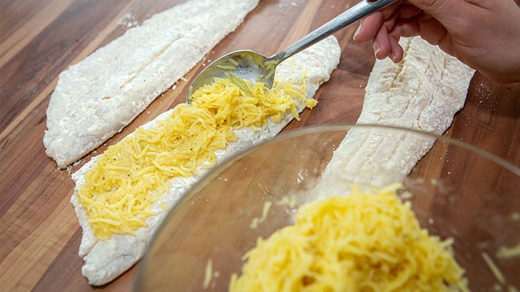 Eine Hand verteilt geriebenen Käse mit einem Löffel auf ausgerollten Teigstreifen, eine Schüssel mit Käse steht im Vordergrund.