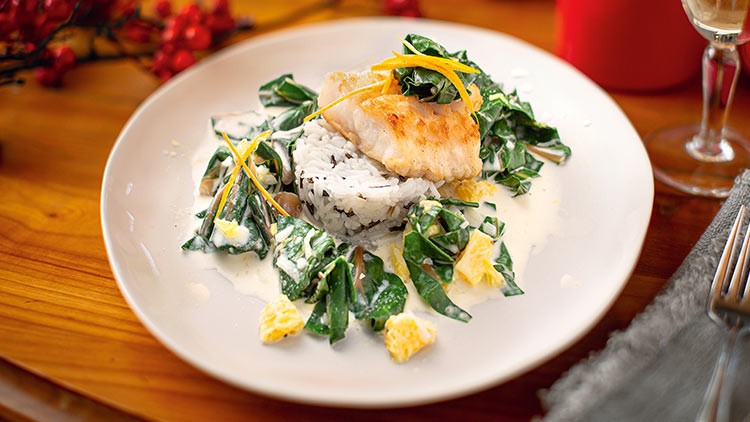Gebratener Fisch mit weißem Reis, Spinat in heller Sauce und Orangenschnitzen auf einem weißen Teller angerichtet.