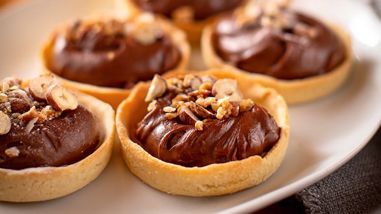 Sechs kleine Tartelettes mit Schokocreme-Füllung und gehackten Haselnüssen auf einem weißen Teller.