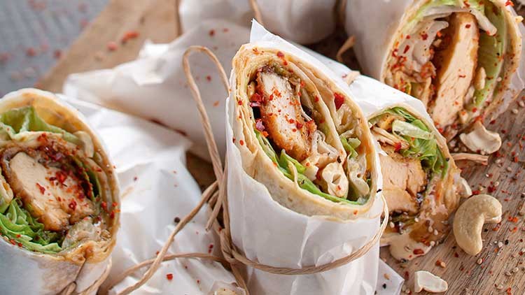 Mehrere Wraps mit Hähnchen, Salat und Gewürzen, teils in Papier gewickelt, liegen auf einem Holzbrett mit Cashewkernen.