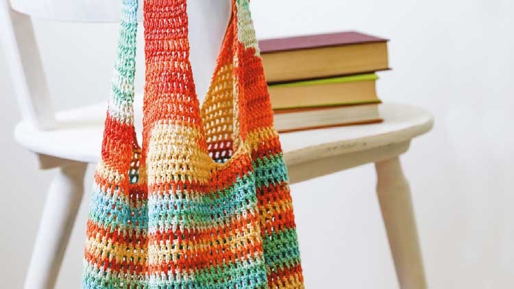 Eine gestrickte Tasche in bunten Streifen hängt an der Lehne eines weißen Stuhls, auf dem Bücher gestapelt liegen.