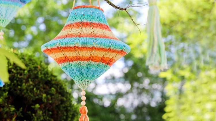 Eine bunt gestrickte, gestreifte Hängelampe mit Quaste hängt an einem Baum vor unscharfem, grünem Hintergrund.