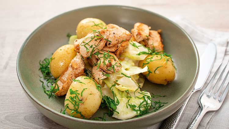 Ein grüner Teller mit kleinen Kartoffeln, gebratenem Fleisch, Weißkohl und frischen Kräutern, daneben Messer und Gabel.