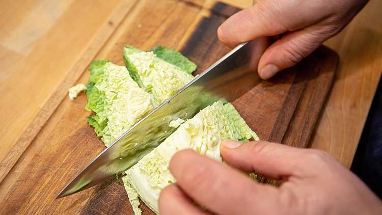 Hände schneiden ein Stück Wirsingkohl mit einem großen Küchenmesser auf einem Holzschneidebrett.