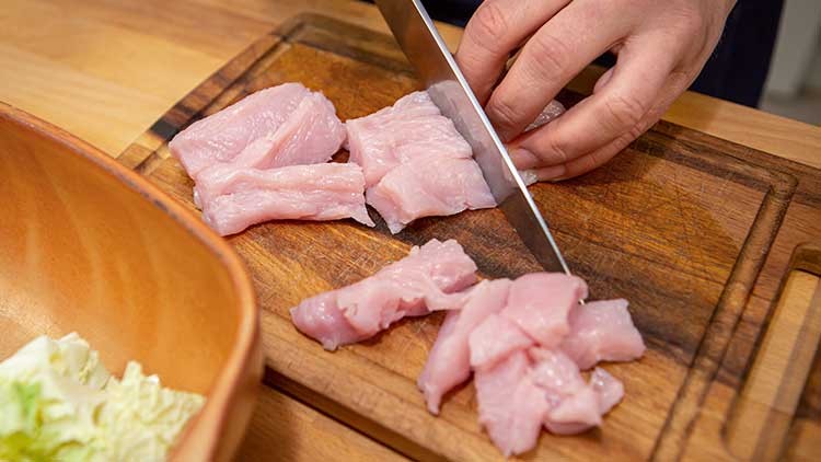 Eine Hand schneidet rohes, hellrosa Hähnchenfleisch auf einem Holzbrett in Stücke, daneben liegt ein Holzschüsselrand.