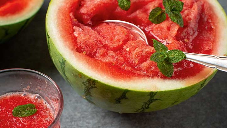 Eine halbierte Wassermelone mit rotem Fruchtfleisch, einem Löffel, Minzblättern und einem Glas mit Wassermelonen-Getränk.