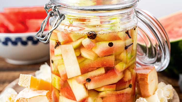 Ein Glas mit eingelegten Wassermelonen-Schalen und Pfefferkörnern steht neben frischer Melone und Rinde auf einem Holztisch.