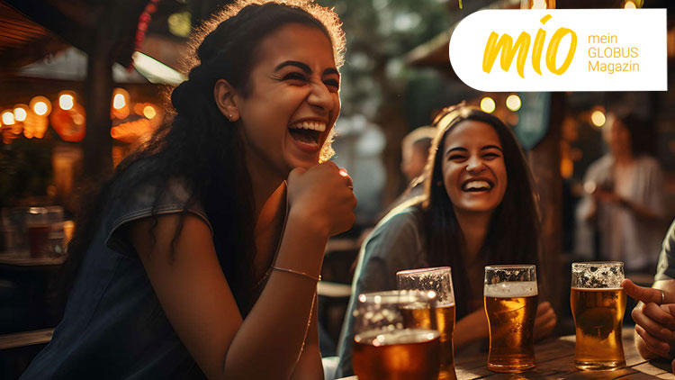 Vier Biergläser stehen auf einem Holztisch in einem gemütlich beleuchteten Außenbereich mit mio mein GLOBUS Magazin-Logo oben links.