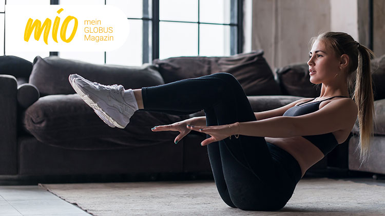 Person in sportlicher Kleidung führt Bauchmuskelübung vor einem Sofa in einem Wohnzimmer aus, mio mein GLOBUS Magazin-Logo links oben.