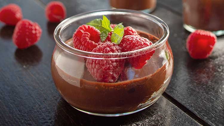 Ein Glas mit Schokoladenmousse, dekoriert mit frischen Himbeeren und grünen Minzblättern, steht auf einem dunklen Holztisch.