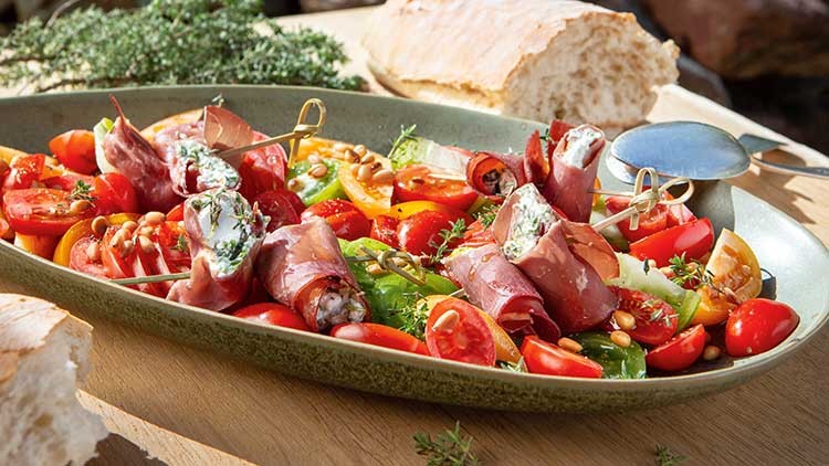Grüne Schale mit gefülltem Schinken, bunter Tomatenauswahl, Pinienkernen und Kräutern, daneben Stücke von hellem Brot.