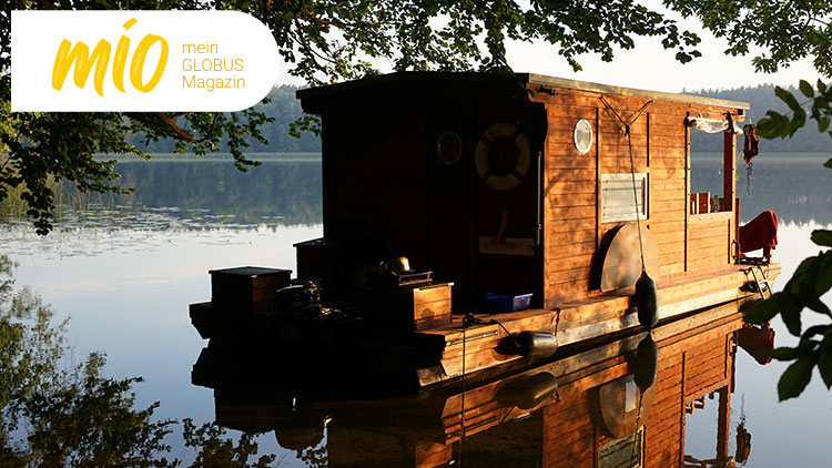 Ein Holzhausboot mit Rettungsring und Fenstern liegt bei Sonnenuntergang an einem ruhigen See, umgeben von Bäumen.