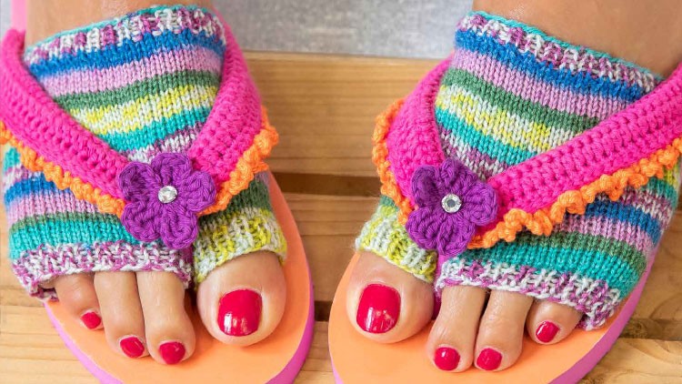 Zehensandalen mit gestricktem, bunt gestreiftem Überzug und lila Blumenverzierung auf lackiertem Holzuntergrund.