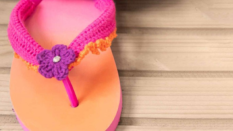 Ein orange-pinker Zehentrenner mit gehäkeltem pink-orangefarbenem Riemen und einer lila Blume liegt auf hellem Holz.