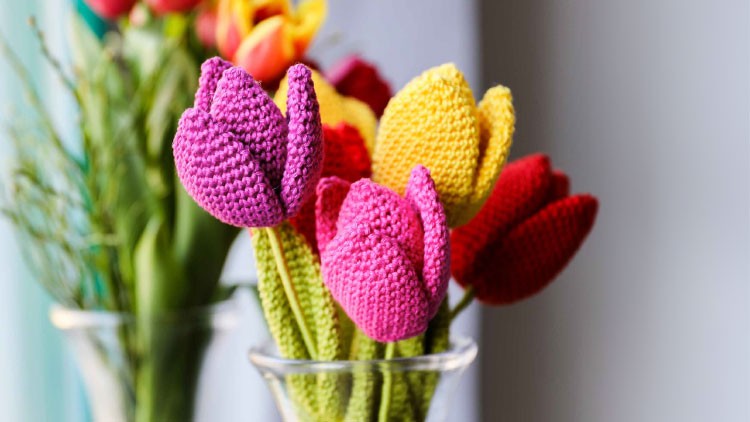 Gehäkelte Tulpen in Gelb, Rot und Pink in durchsichtigen Gläsern vor einem unscharfen Hintergrund mit weiteren Blumen.