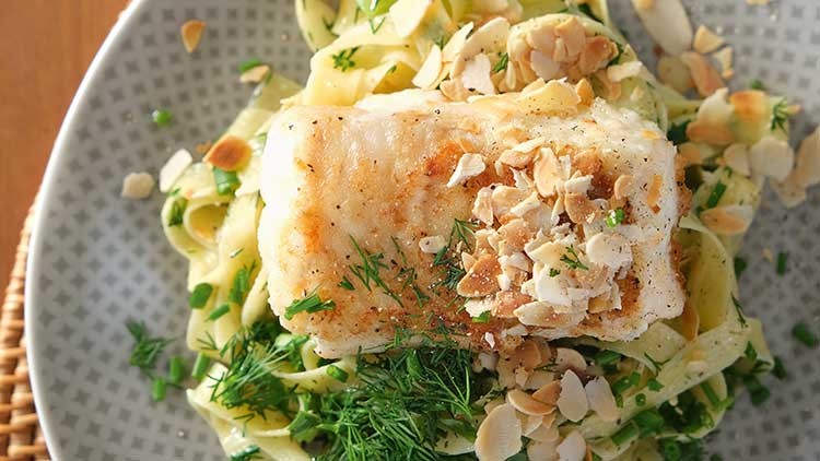 Gebratener Fisch mit gehobelten Mandeln auf einem Bett aus Bandnudeln, frischen Kräutern und Dill in einem grauen Teller.