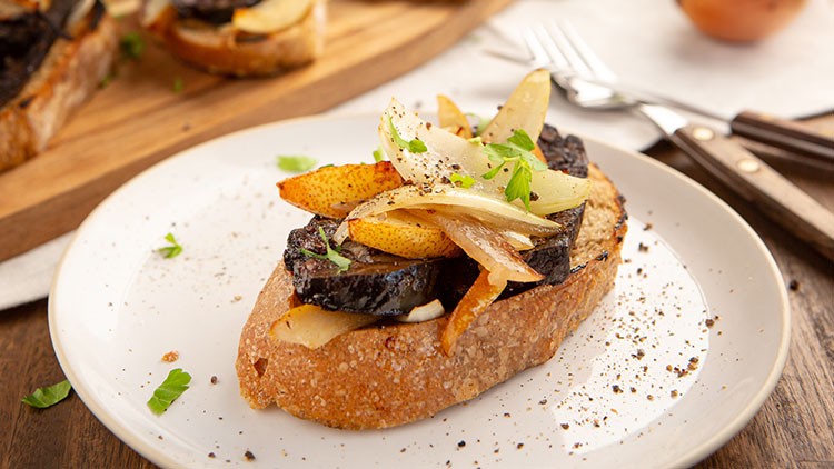 Ein Stück Brot belegt mit angebratener Blutwurst, karamellisierten Zwiebeln, Birnenscheiben und Kräutern auf einem weißen Teller.