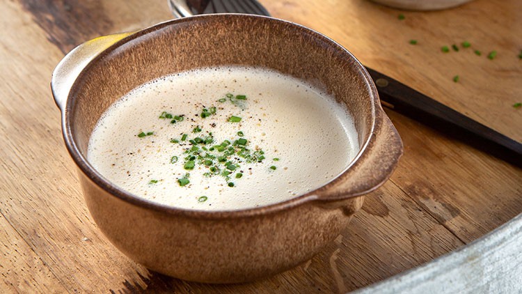 Schaumige Suppe in brauner Schale mit Schnittlauchstücken auf einem rustikalen Holztisch, daneben liegt ein Löffel.