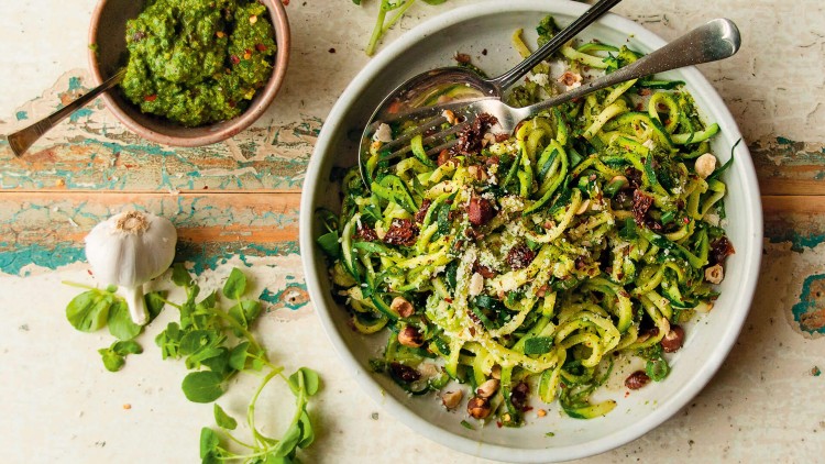 Auf einem Teller sind Zucchini-Spaghetti mit grünem Pesto, Nüssen und getrockneten Tomaten, daneben eine Schale mit Pesto.