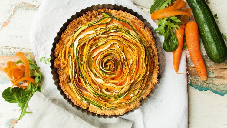 Runde Gemüse-Tarte mit spiralförmig angeordneten Zucchini- und Karottenstreifen auf einem weißen Tuch, daneben frisches Gemüse.