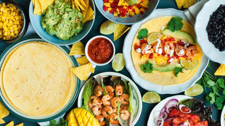 Bunte Auswahl an mexikanischen Speisen wie Tacos, Guacamole, Salsa, Garnelen, Tortillas, Nachos, Mango, Mais, Bohnen und Salat.