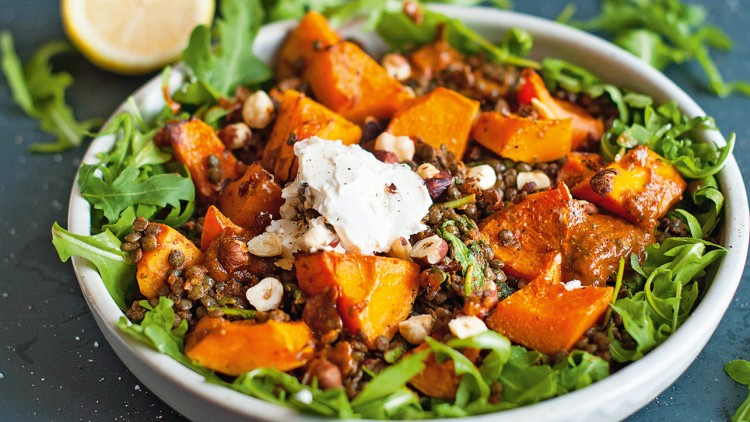 Salat mit Rucola, gebackenen Kürbisstücken, Linsen, Nüssen und einem Klecks Frischkäse auf dunklem Untergrund.