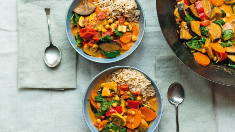 Zwei Schalen mit Gemüsecurry und Reis stehen auf einem hellen Tuch neben zwei Löffeln, daneben eine große Pfanne mit Curry.