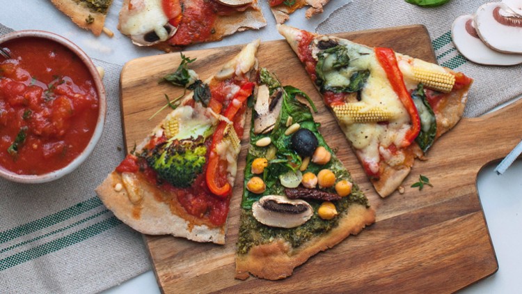 Eine bunte Pizza mit Brokkoli, Paprika, Mais und Oliven auf Holzbrett, daneben Tomatensoße, frische Basilikumblätter und Pilze.