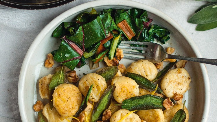 Gebratene Gnocchi mit Salbei, Zitrone und Walnüssen in einer Schale, daneben Mangold und eine Gabel auf hellen Stoffen.