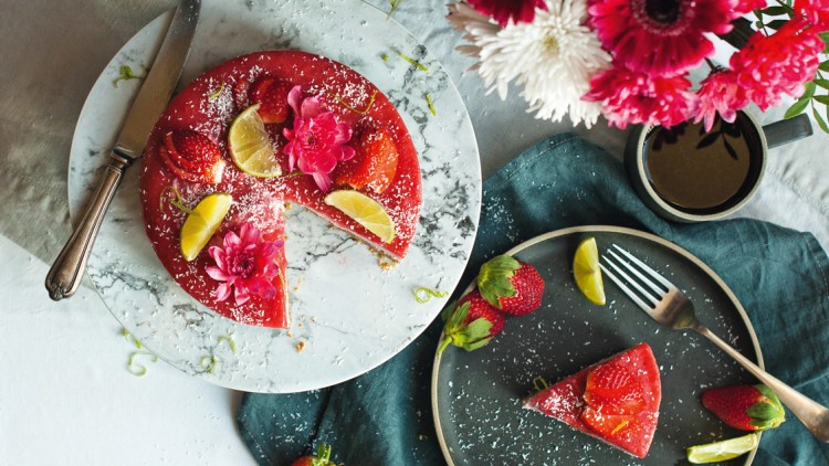 Roter Kuchen mit Erdbeeren, Limettenstücken und rosa Blüten auf Marmorplatte, daneben ein Kuchenteller und ein Blumenstrauß.