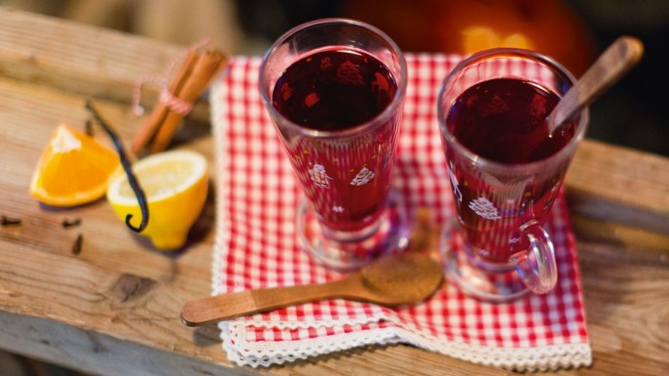 Zwei Gläser mit rotem heißen Getränk auf rot-weiß-kariertem Tuch, daneben Zimt, Vanilleschote, Orange und Zitrone.