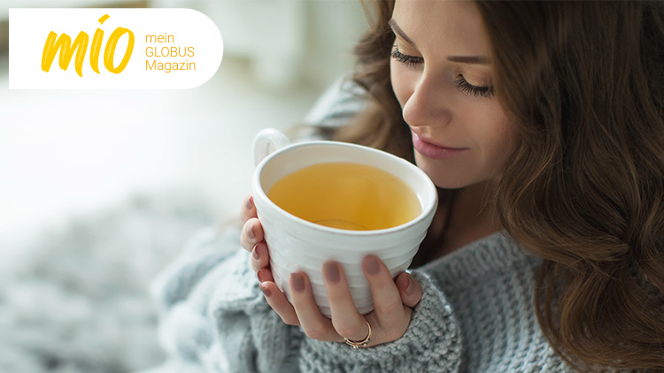 Eine Person hält eine weiße Teetasse mit hellem Tee, links oben ist der Schriftzug „mio mein GLOBUS Magazin“ in gelb und orange.