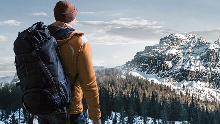 Person mit Rucksack blickt in verschneite Berglandschaft mit Tannen und großem Felsen, leichter Himmel mit Wolken.