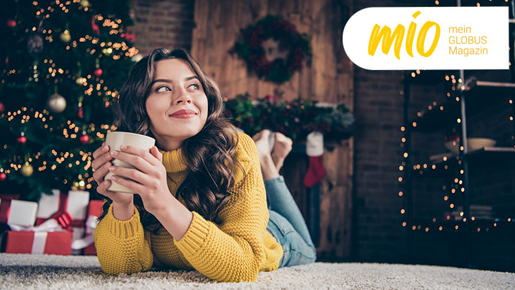 Person in gelbem Pullover hält eine Tasse vor einem weihnachtlich dekorierten Baum, daneben steht das mio mein GLOBUS Magazin Logo.