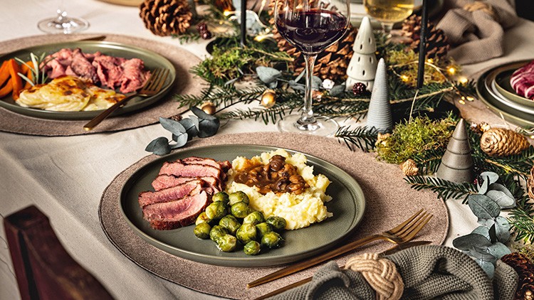 Festlich gedeckter Tisch mit Teller, Rosenkohl, Kartoffelpüree, Bratenscheiben und Soße, dazu Dekoration und Rotweinglas.